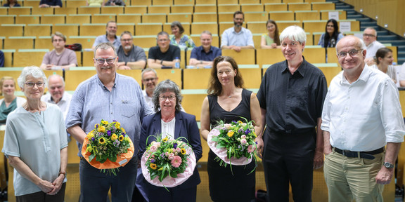 Eine Gruppe von sechs Personen, darunter die drei neuen Prorektor*innen, halten bunte Blumensträuße, steht in der vorderen Reihe eines Hörsaals. Sie lächeln in die Kamera.